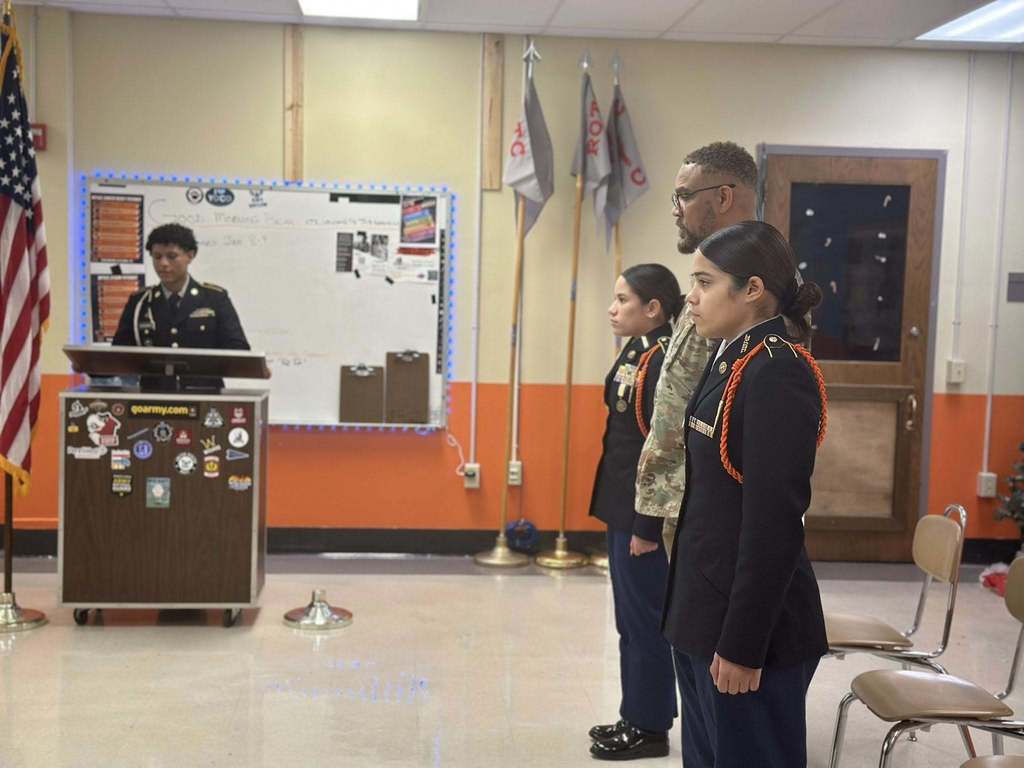 Three uniformed individuals are standing in a line, facing an instructor at a podium with an American flag. The room has military decor with flags and a colorful bulletin board.