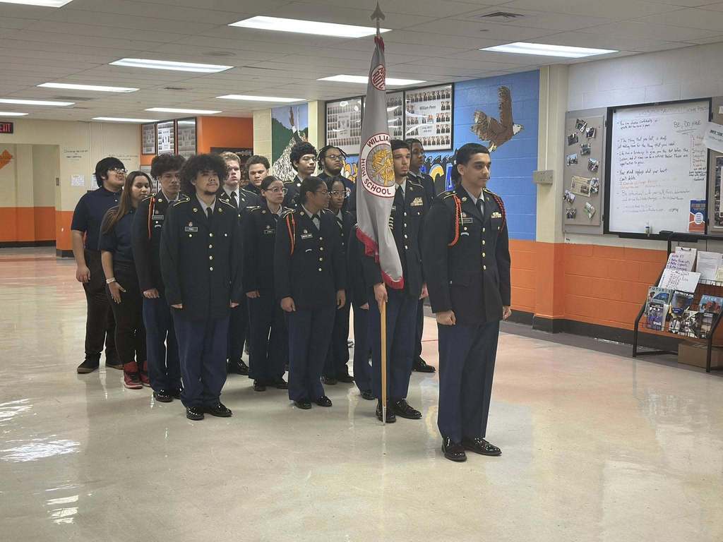 A group of uniformed students are standing in formation in a school classroom. One student is holding a flag labeled "High School." The mood is formal and organized. Posters and information are displayed on the walls.