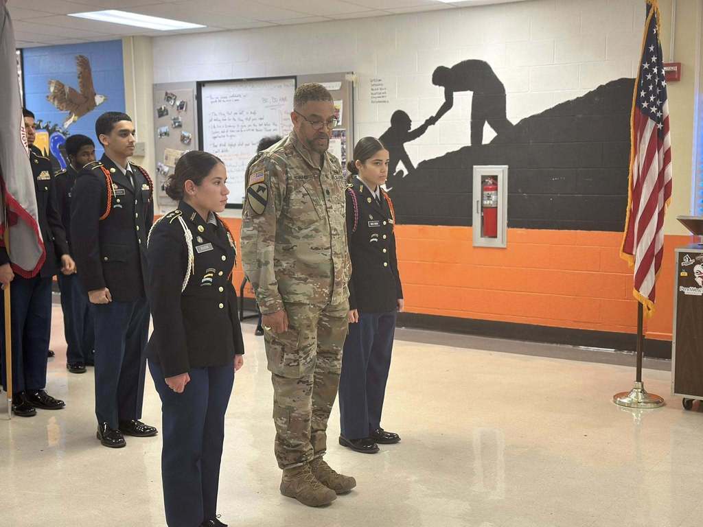 A group is standing solemnly in a school classroom, including people in military uniforms. A mural of one person helping another is on the wall.