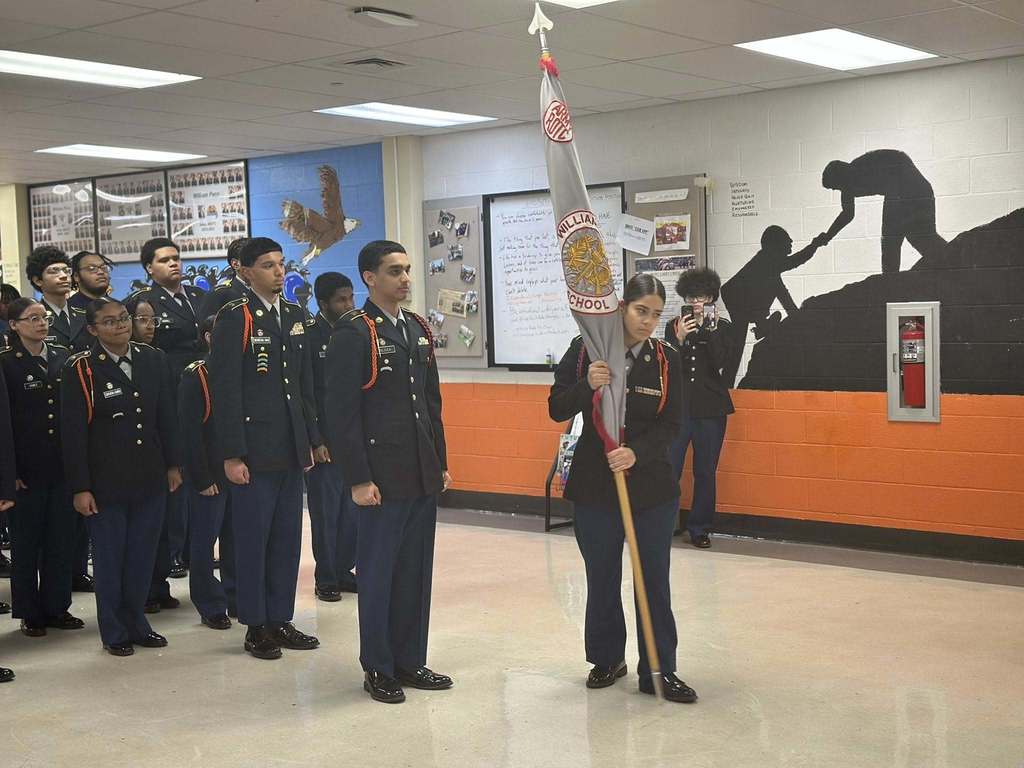 A group of students in military uniforms are standing in formation in a school classroom. One student is holding a flag. A mural on the wall is depicting a person helping another climb.