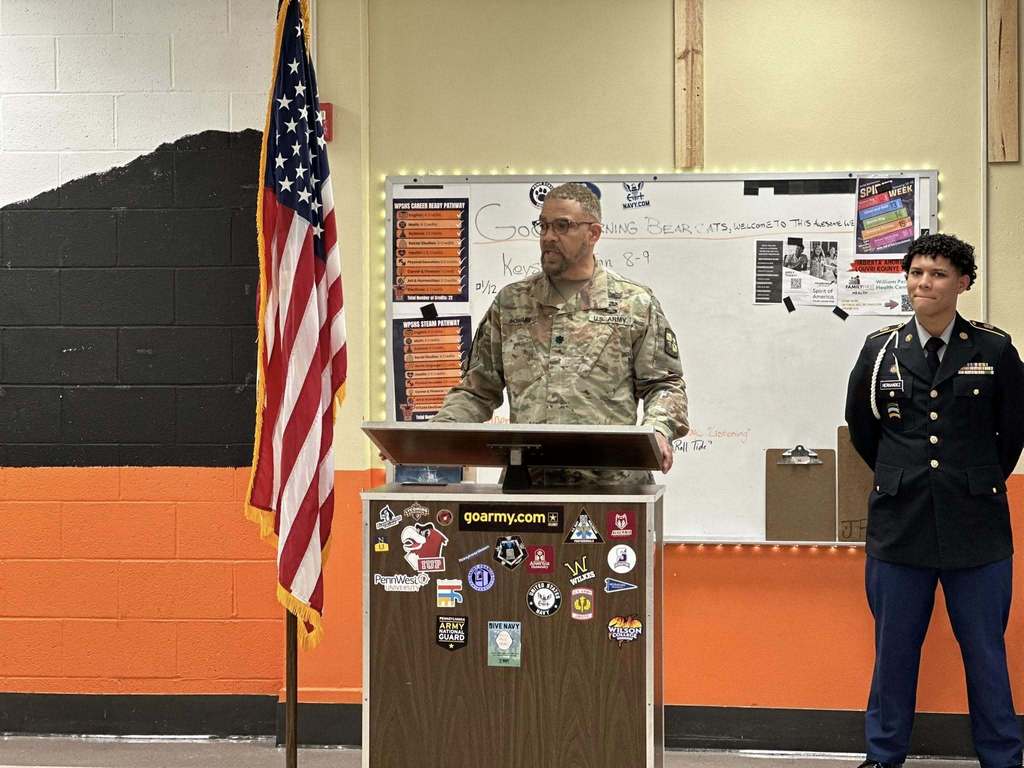 A man in military uniform is speaking at a podium adorned with various stickers. An American flag is next to his left and a uniformed individual is standing to his right, near a whiteboard.