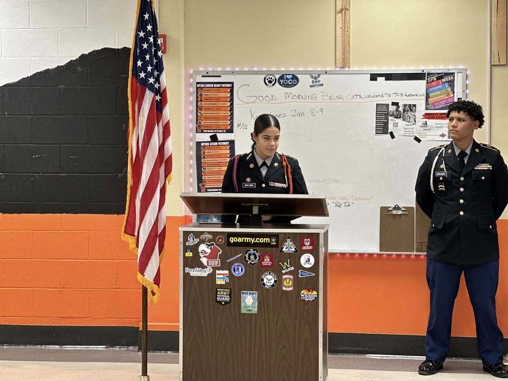 A young woman in a military uniform is standing at a lectern with an American flag beside her. A young man in uniform is standing to the right of her. A whiteboard with notes and a background of orange and black walls are visible.