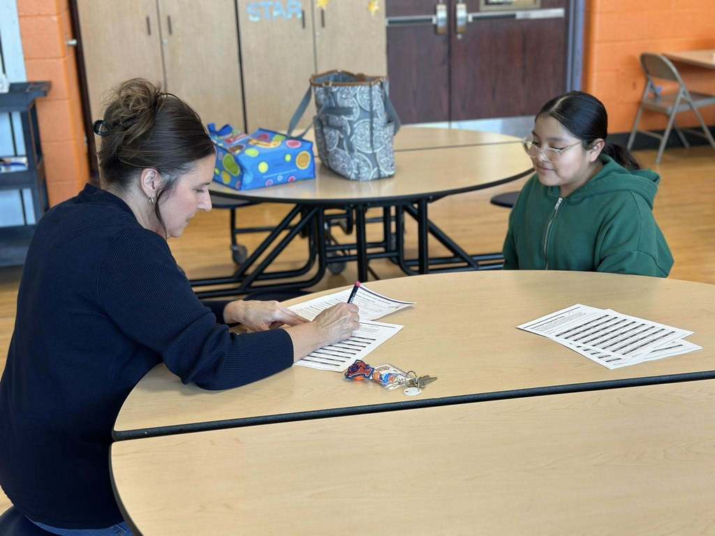 A district staff member and a student are sitting across from each other at a round table in a school cafeteria. The district staff member is writing on papers. 