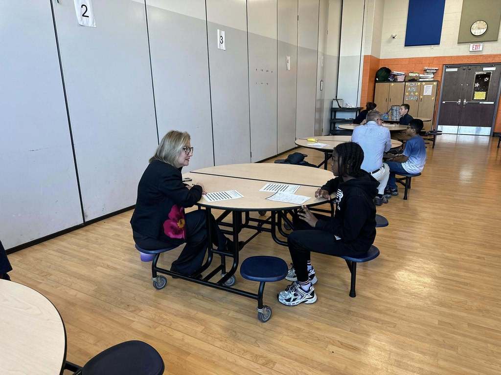 A district staff member and a student are sitting across from each other at a round table in a school cafeteria, engaging in discussion. Other people can be seen in the background.