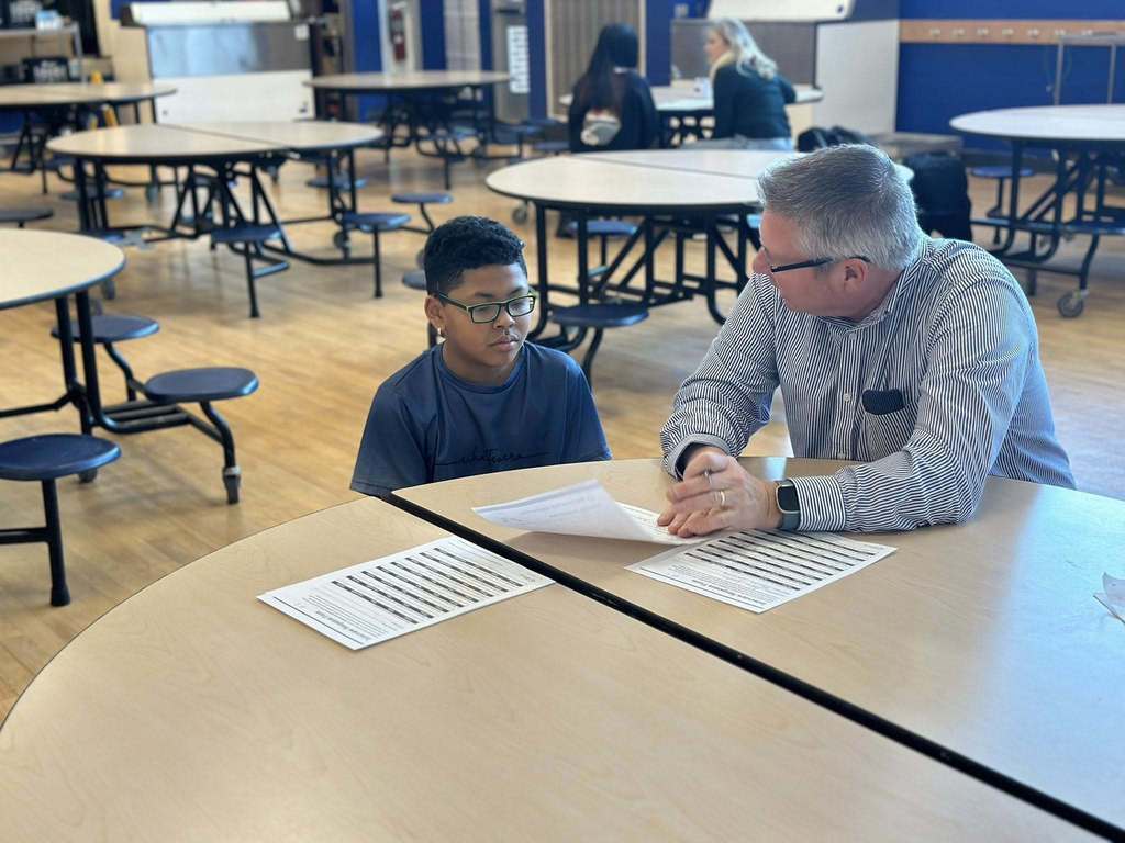 A district staff member is engaging in discussion with a student in a school cafeteria. Other people can be seen in the background.