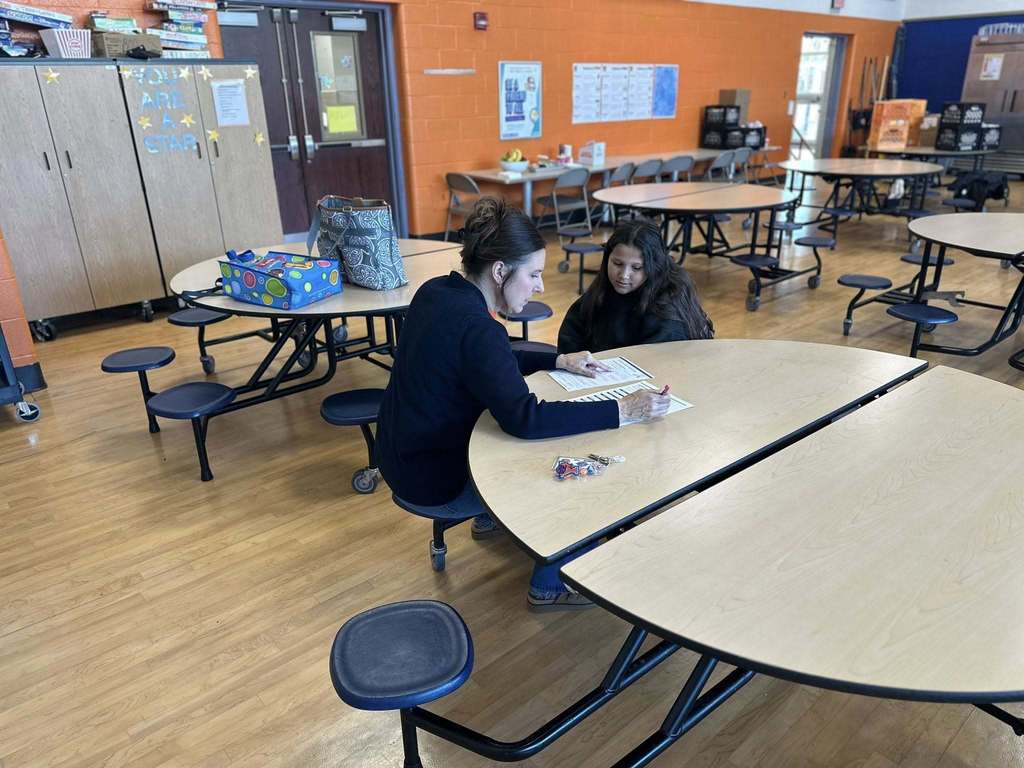 A district staff member and a student are sitting at a round table in a school cafeteria, engaging in discussion. 