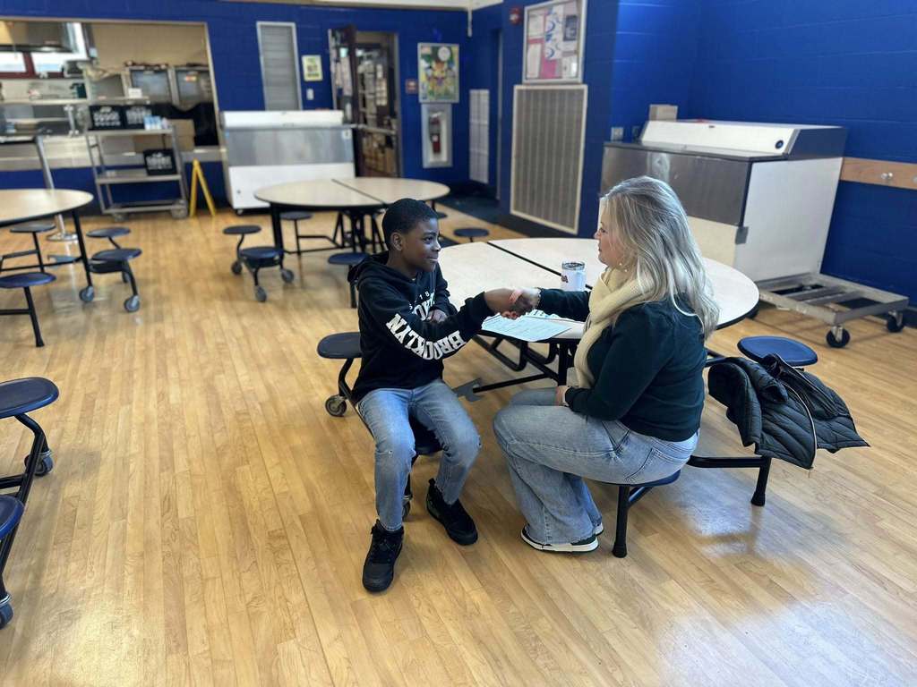 A district staff member and a student are shaking hands while sitting at a round table in a school cafeteria.