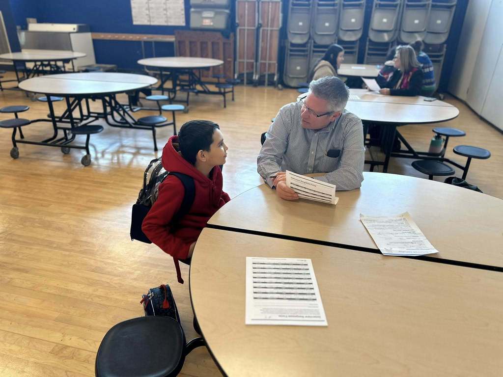 A district staff member and a student are sitting at a round table in a school cafeteria, engaging in discussion. Other people can be seen in the background.