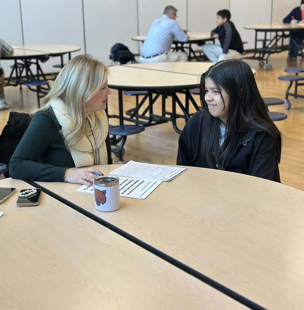 A district staff member and a student are sitting at a roundtable in a school cafeteria, engaging in discussion. Other people can be seen in the background.