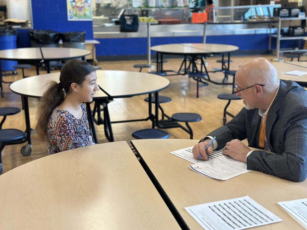 A district staff member and a student are sitting across from each other at a round table in a school cafeteria, engaging in discussion. Other people can be seen in the background.