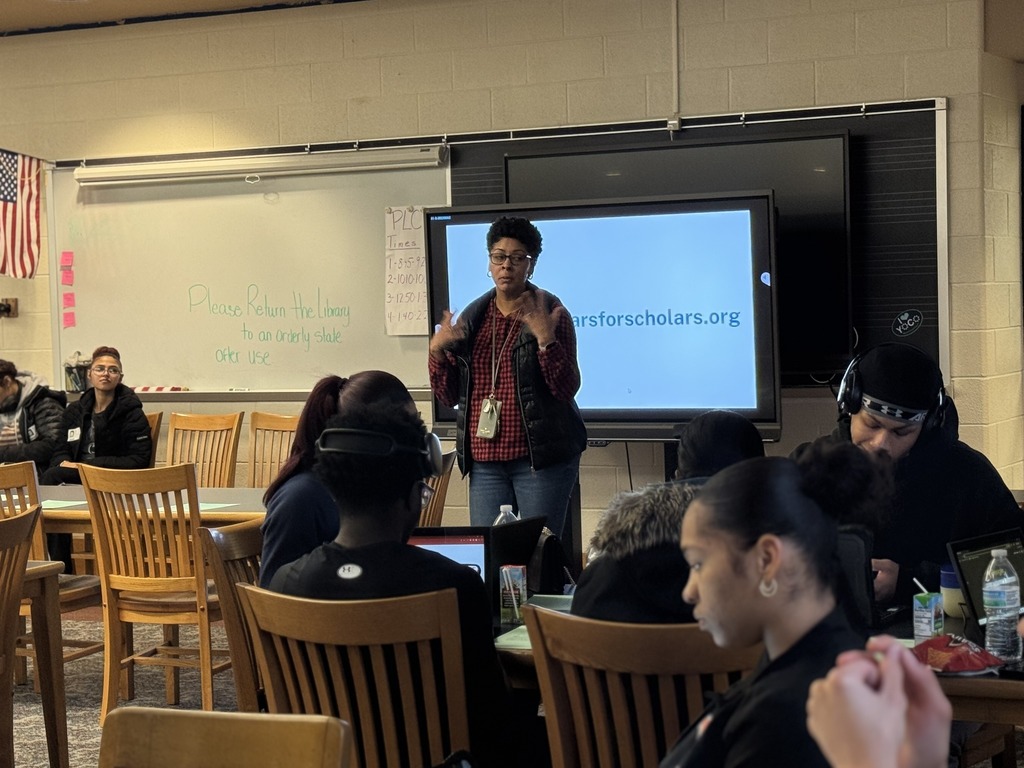 A district staff member is standing in front of a screen, speaking to a group of students who are seated at tables in a school library.
