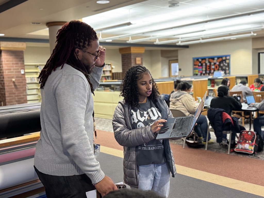 A district staff member and a student are standing in a school library, engaging in discussion while looking at a laptop. Other people can be seen sitting at tables in the background.