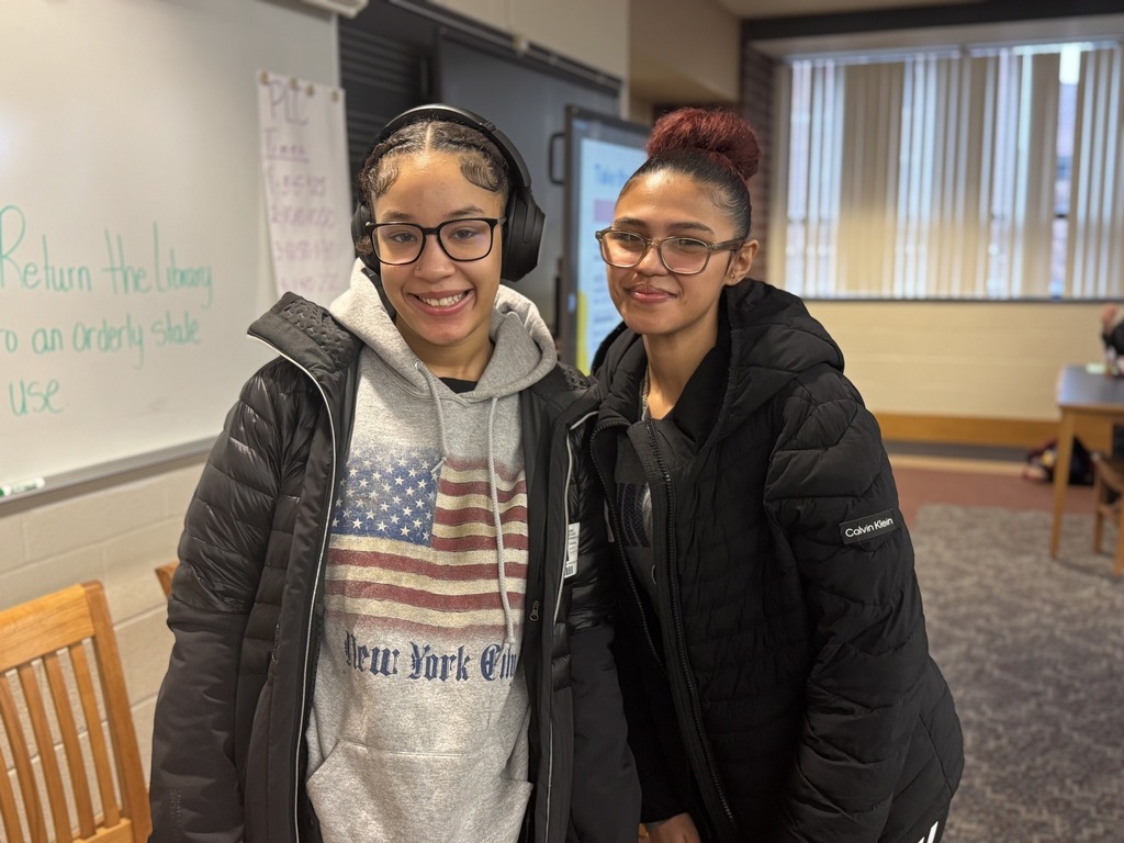 Two students are standing in a school library near a whiteboard. One of the students is wearing headphones.