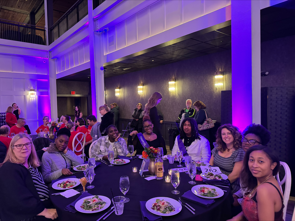 A group of district staff members, former students, and current students are sitting around a table at an elegant event. Plates of salad, candles, and drinks can be seen on the table.