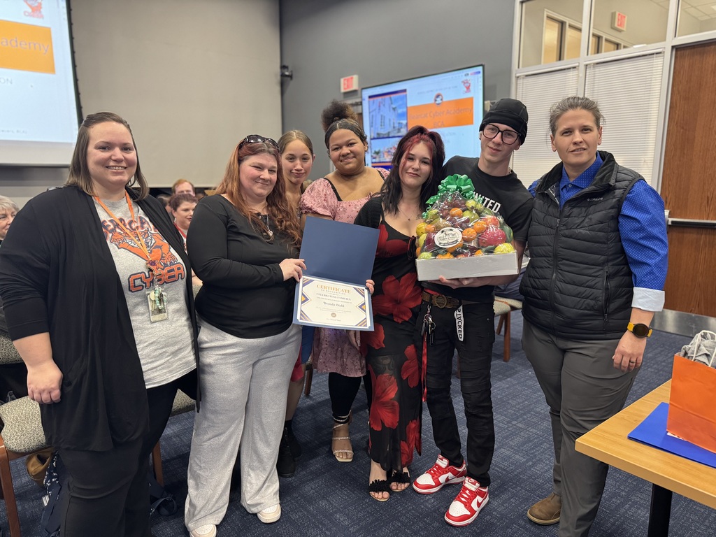 A group of six people including district staff members and a district family are standing in a conference room. A woman is holding a certificate and a young man is holding a colorful fruit basket. An audience of people can be seen in the background.