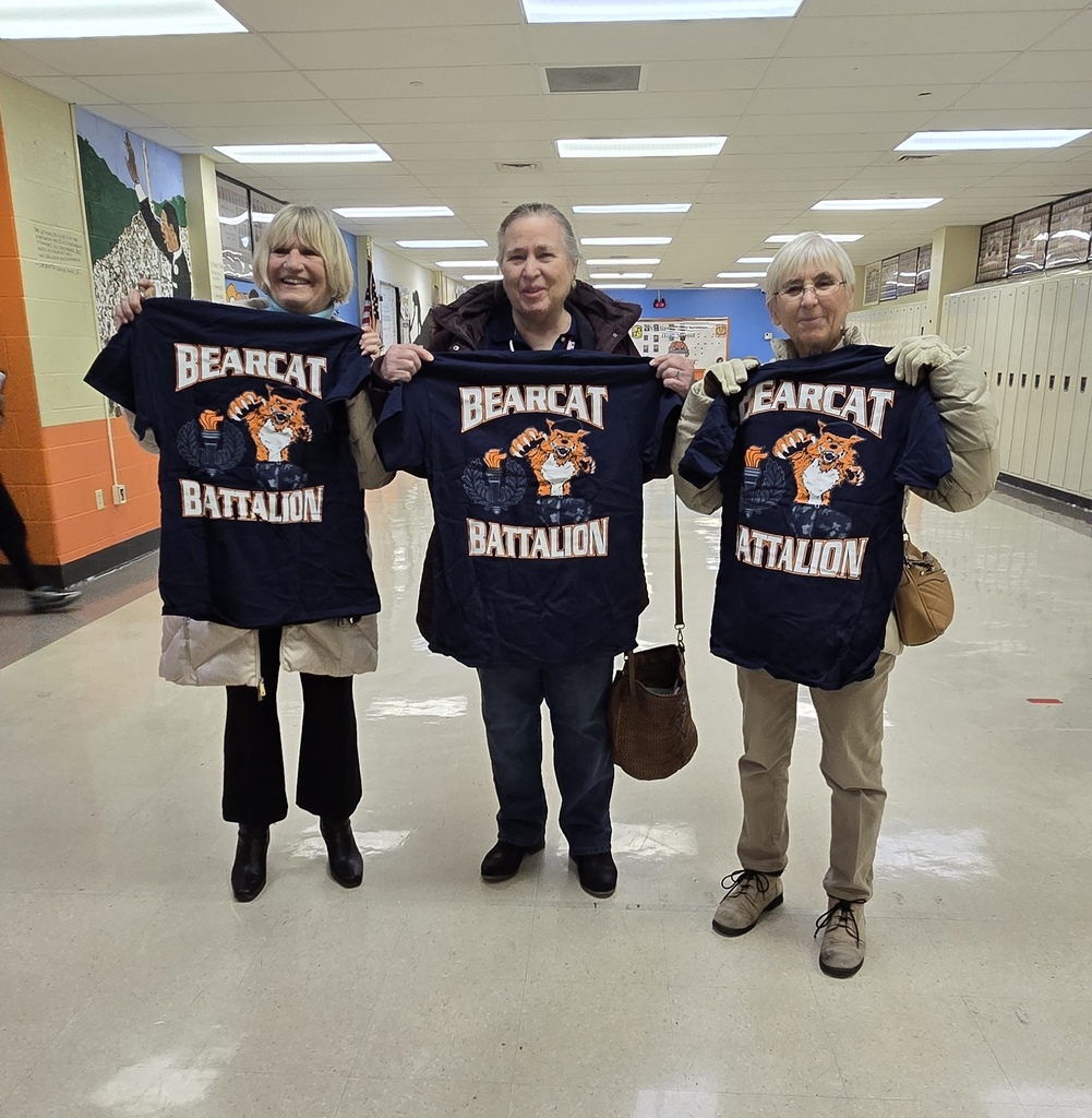 A group of three women are standing in a school classroom, each of them holding a navy "Bearcat Battalion" T-shirt with a Bearcat graphic.