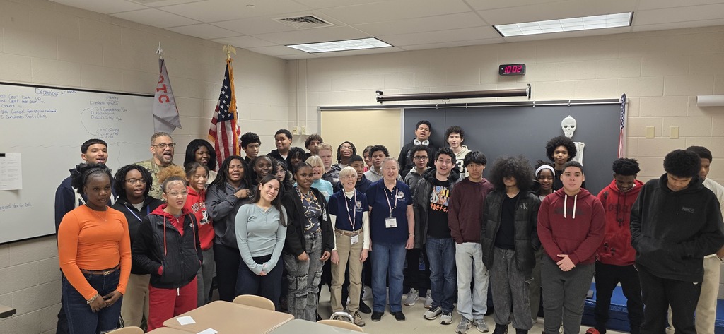 A group of students, a district staff member, and local community members are standing in front of a whiteboard and an American flag in a school classroom. 