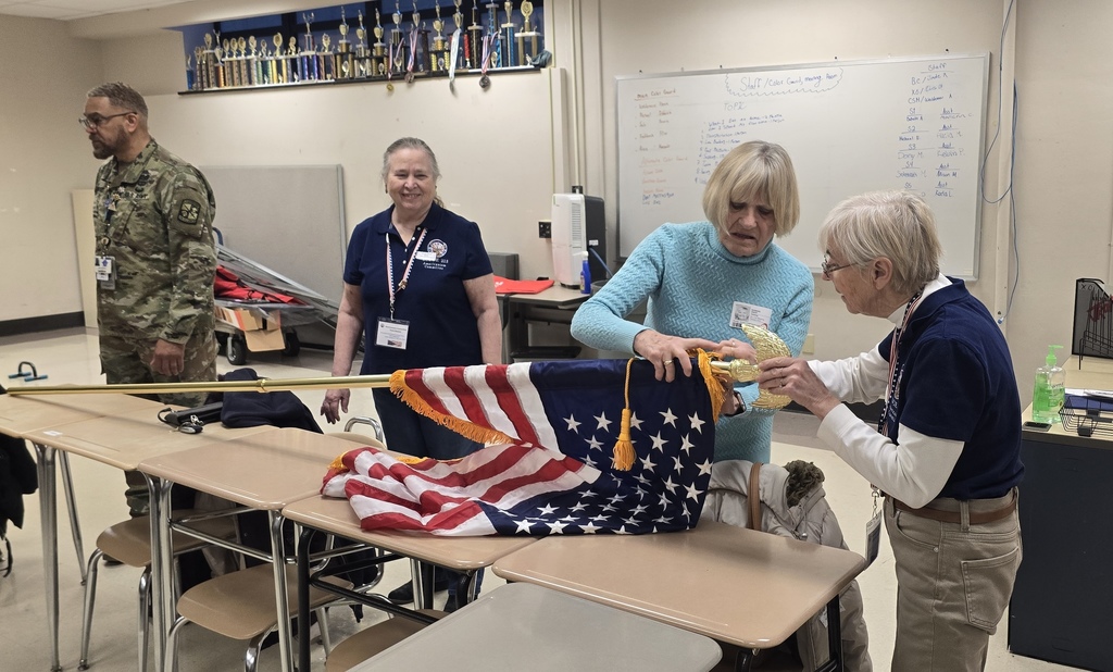 Two women are assembling a U.S. flag, while standing in a school classroom surrounded by desks and a whiteboard. A district staff member in military uniform can be seen standing nearby in the background.