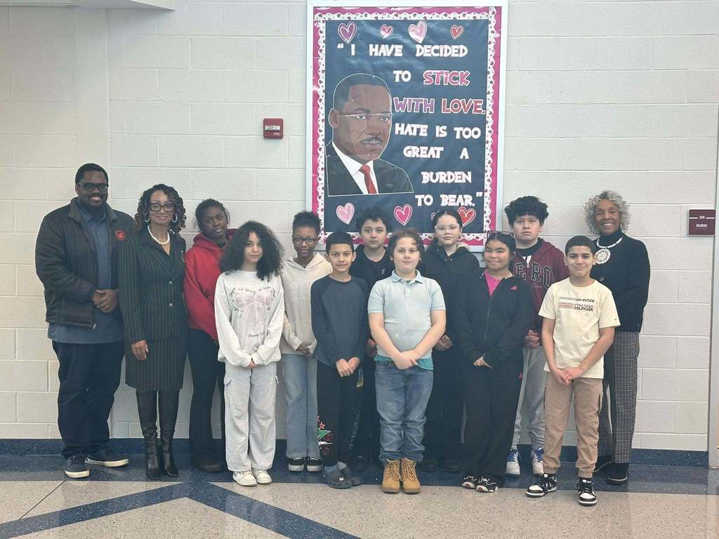 A group of ten students, district administration, and Representative Carol Hill-Evans are standing in a school hallway in front of a colorful mural of Martin Luther King Jr. with the quote, "I have decided to stick with love. Hate is too great a burden to bear." 