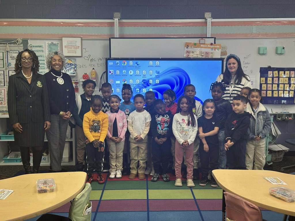 A group of students, district administration, a district staff member and Representative Carol Hill-Evans are standing on a colorful carpet in a school classroom. Behind them is a large screen displaying a desktop.