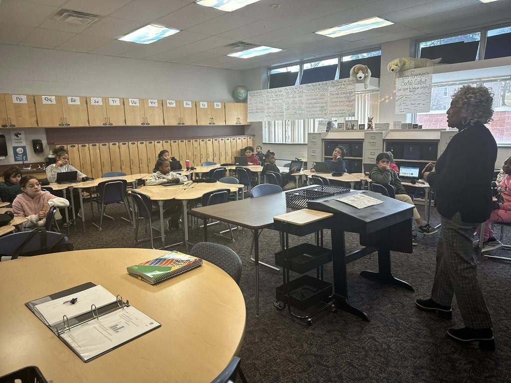 Representative Carol Hill-Evans is standing near a desk, speaking to a group of students who are seated at desks with laptops in a school classroom.