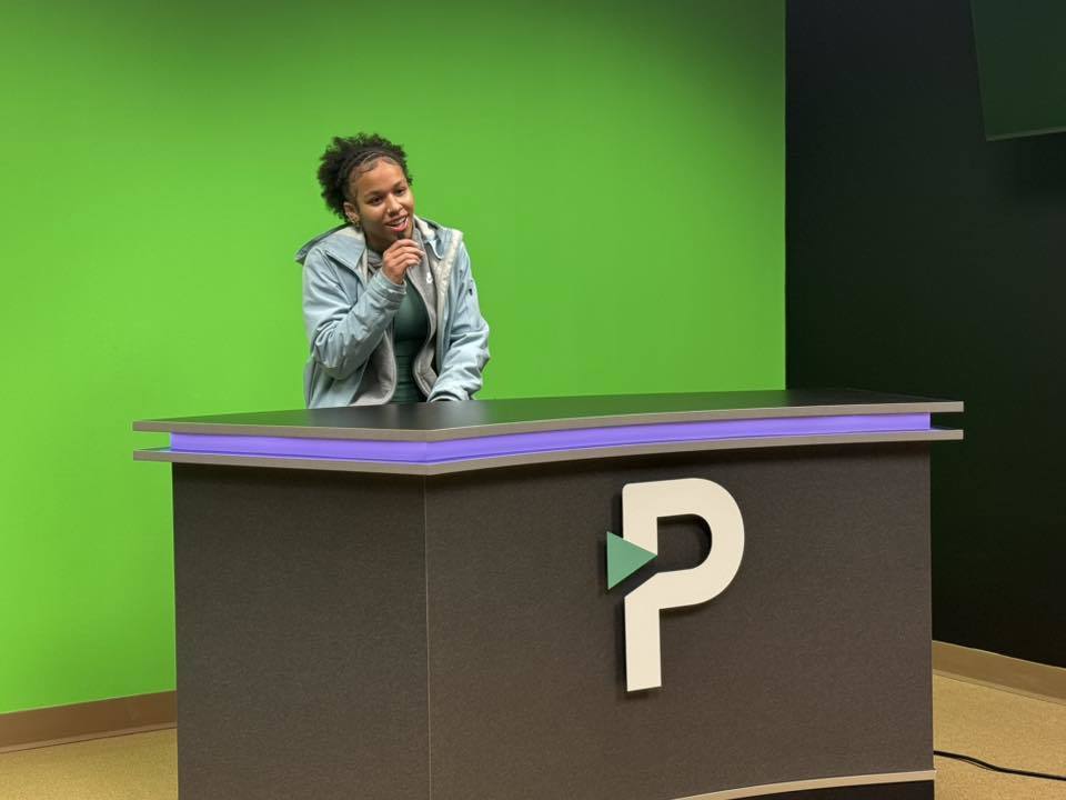 A student is sitting behind a news desk in front a green screen backdrop, holding a microphone. The desk has a large "P" logo.