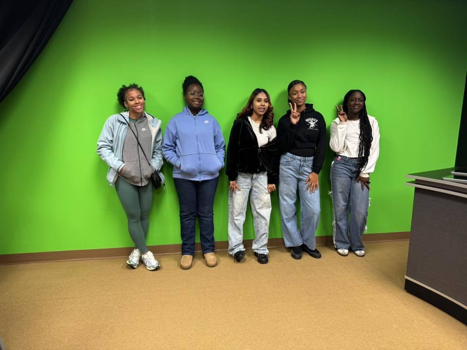 Five young women are standing side by side against a bright green backdrop, smiling and posing. Two of the young women are holding up peace signs.