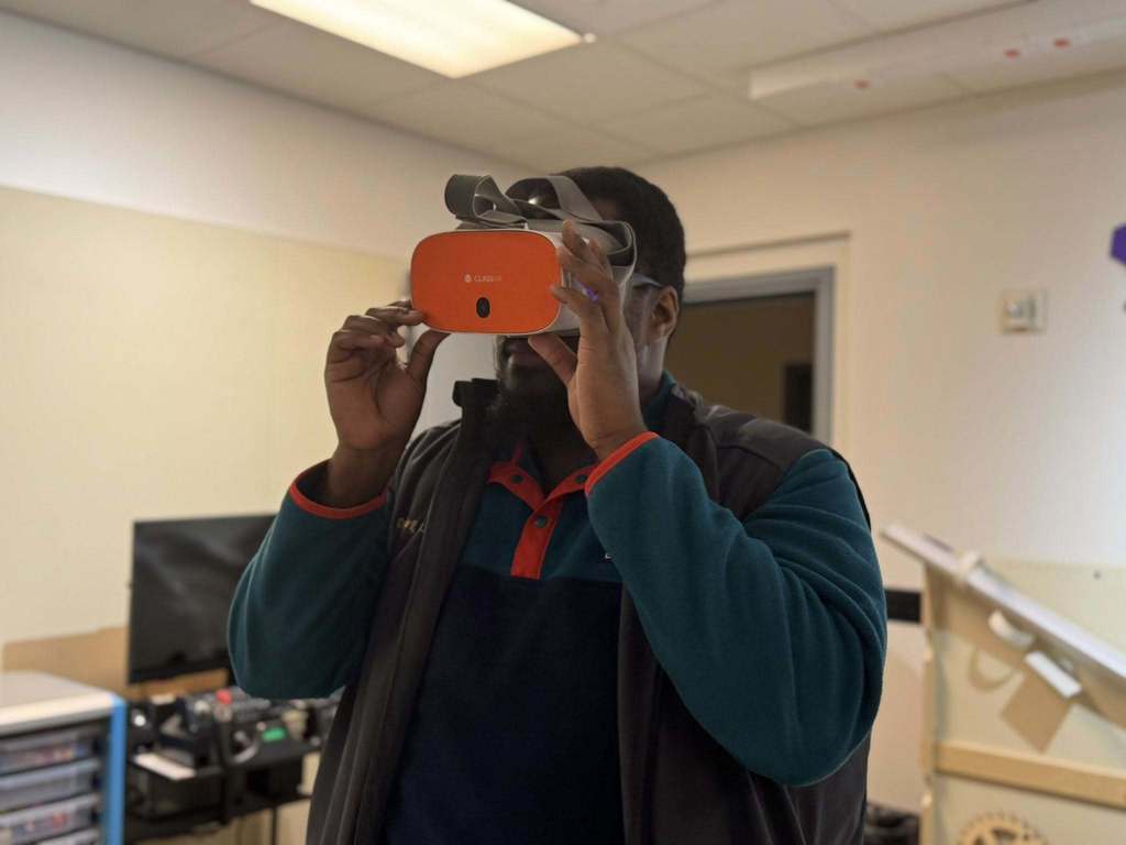 District administration is wearing an orange virtual reality headset while standing in an office area with computers around.