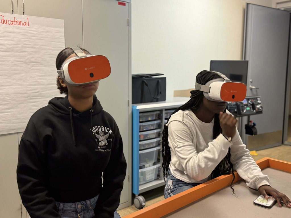 Two students are sitting at a table wearing VR headsets in a classroom setting. One of the students appears to be interacting with the device. A chart can be seen in the background.