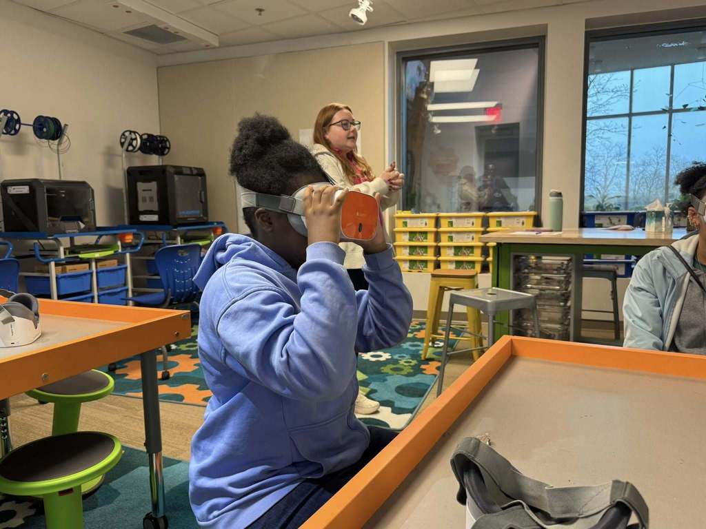 A student is wearing an orange virtual reality headset in a classroom area. An adult can be seen standing nearby in the background and another student can be seen sitting nearby.