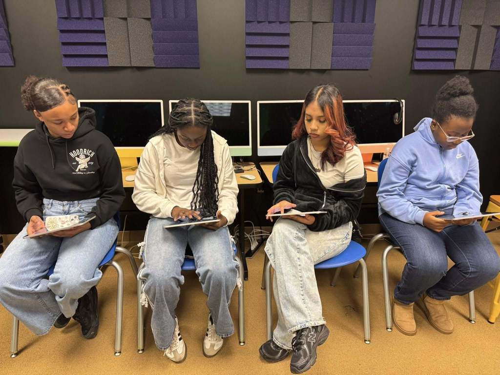 Four young students are sitting in a computer lab, working on tablets. Behind them are monitors and soundproof panels.