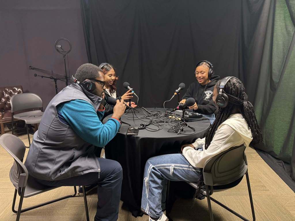District administration and a group of three students are sitting around a table in a recording studio, wearing headphones and speaking into microphones. 
