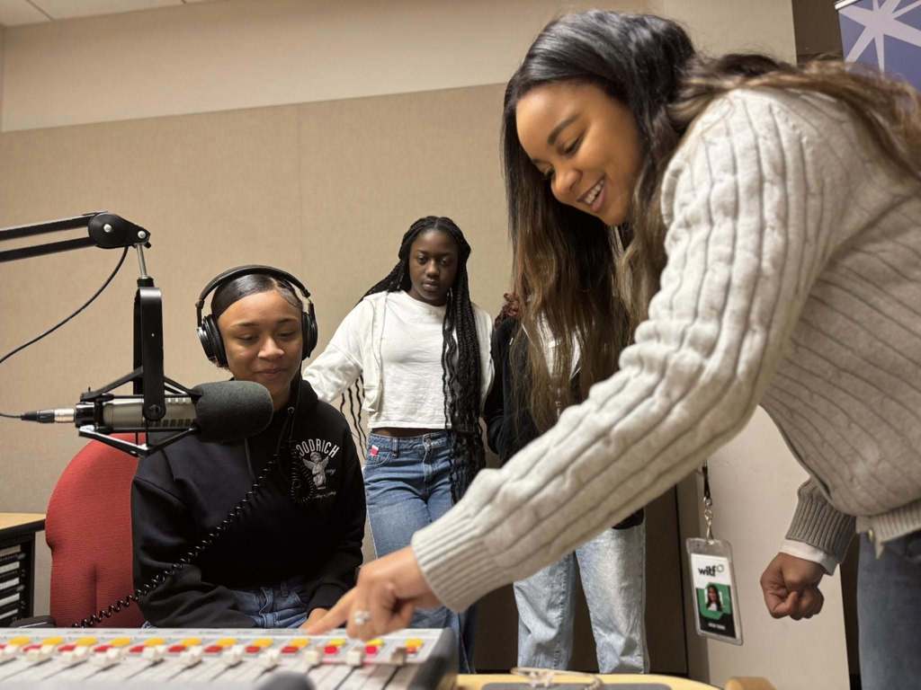 A woman is adjusting a soundboard in a radio studio. A student is sitting nearby wearing headphones, while two other students are watching attentively. 
