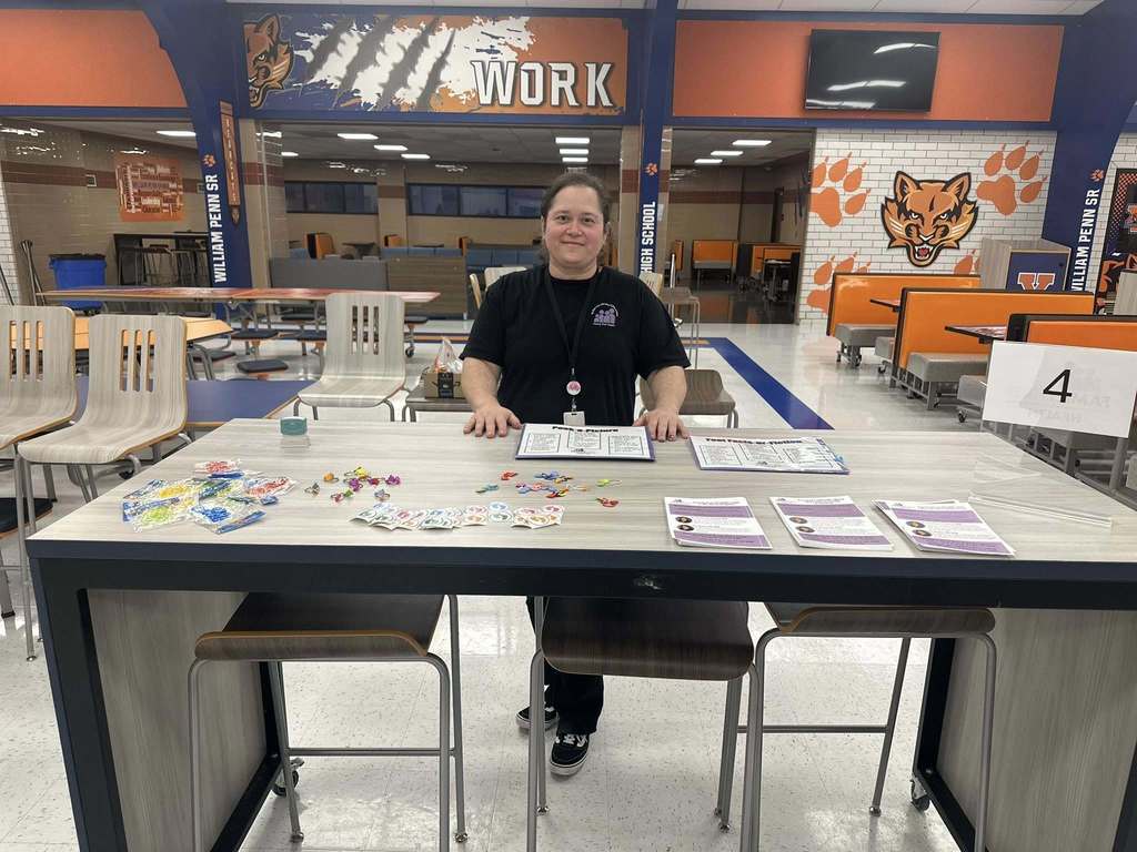 A person is standing at a table in a school cafeteria. The table is displaying colorful trinkets and papers.