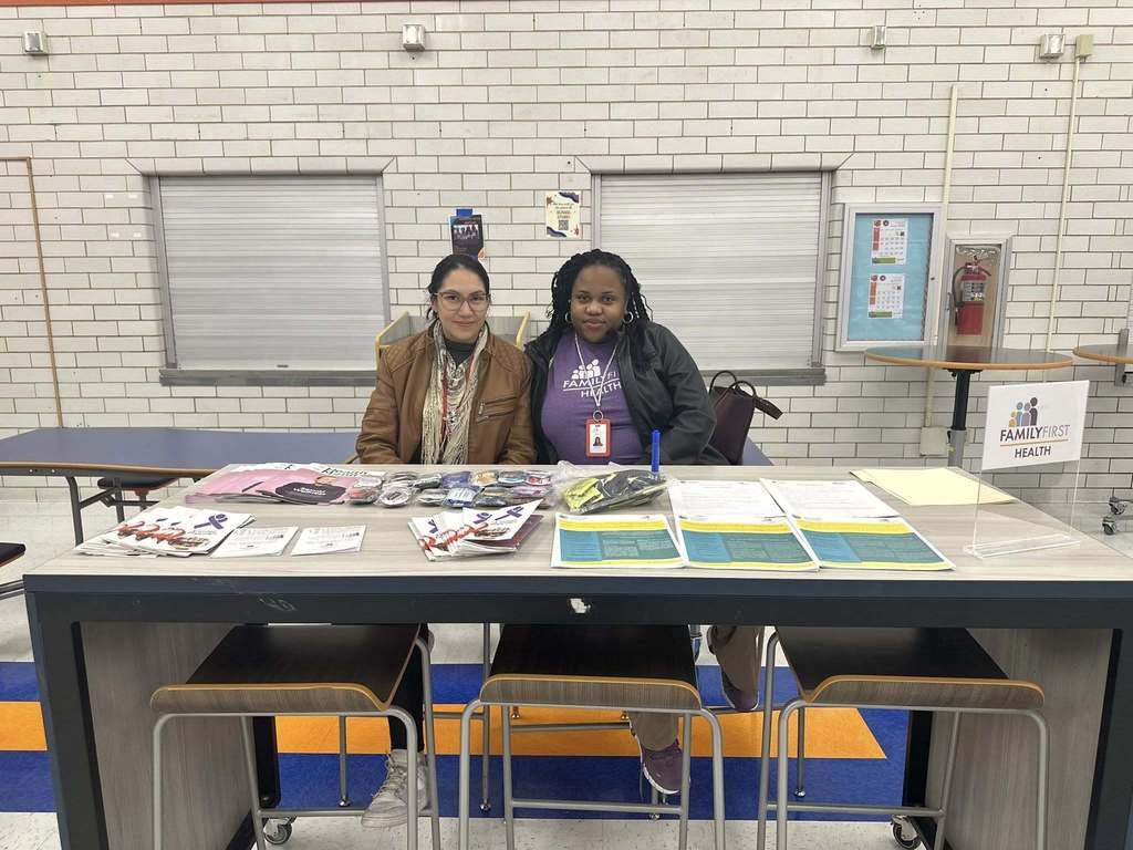 Two district staff members are sitting at a table in a school cafeteria. The table is displaying brochures, flyers, and small items. A "Family First Health" sign is visible.