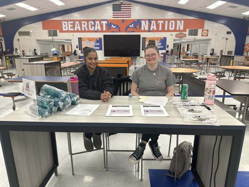 Two district staff members are sitting at a table in a school cafeteria labeled "Bearcat Nation." The table has flyers, bottles, and giveaways.