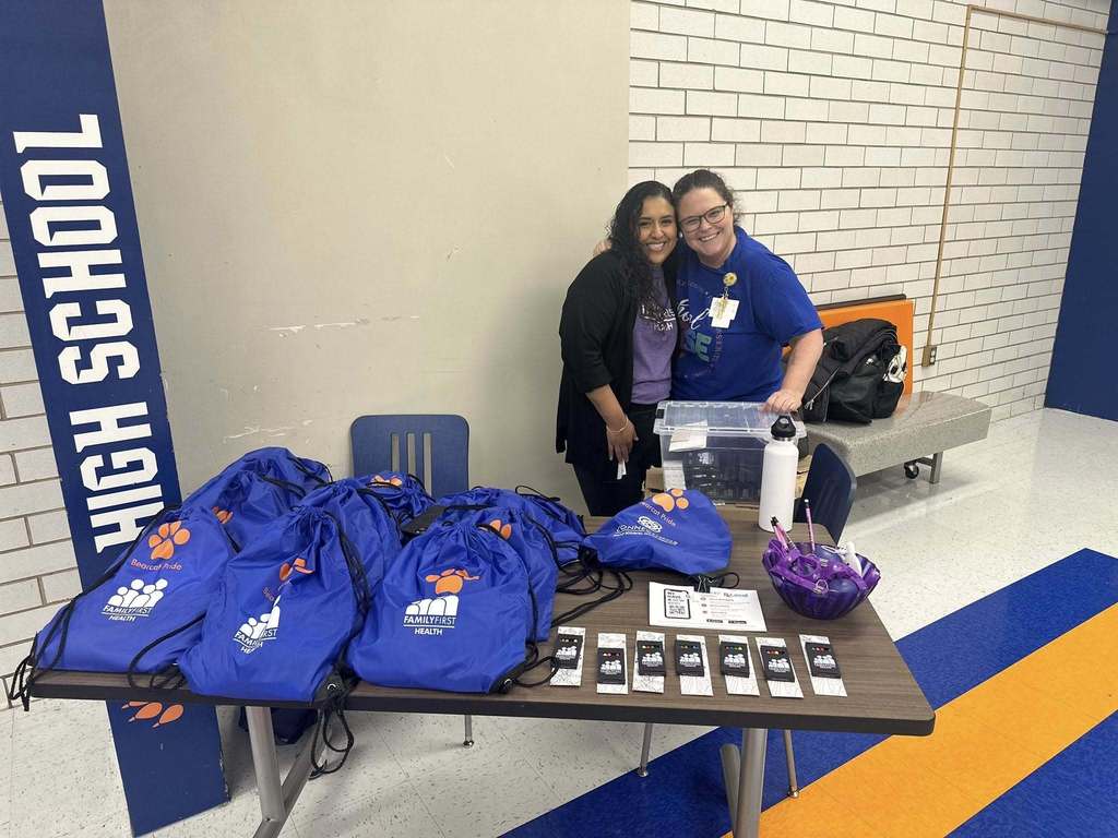 Two district staff members are standing at a table in a school cafeteria with blue drawstring bags labeled "Family First Health."