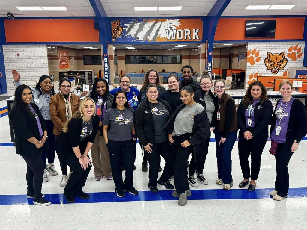 A group of 16 Family First Health employees are standing in a school cafeteria with vivid orange and blue decor. Some of them are wearing branded shirts.