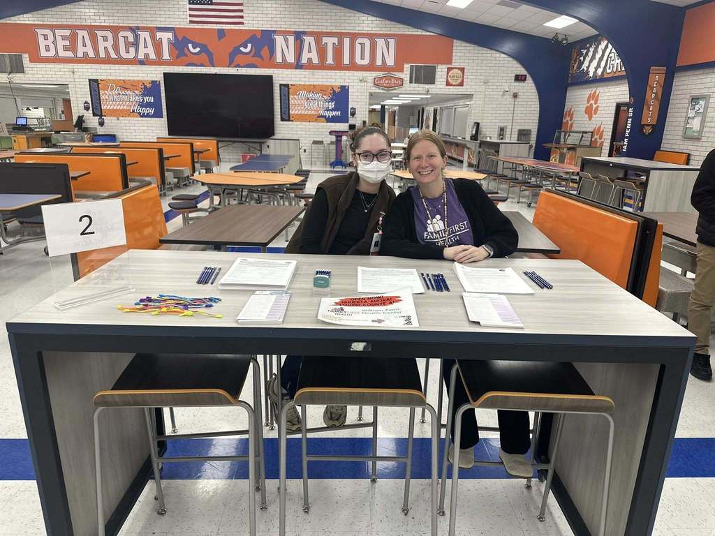 Two district staff members are sitting at a table in a school cafeteria with "Bearcat Nation" on the wall. The table has papers, pens, and a number "2" sign.