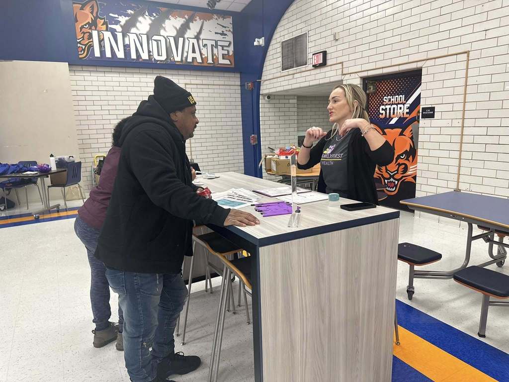 A district staff member and a man are engaging in conversation at a table in a school cafeteria. The backdrop features a white brick wall with "Innovate" in bold letters.