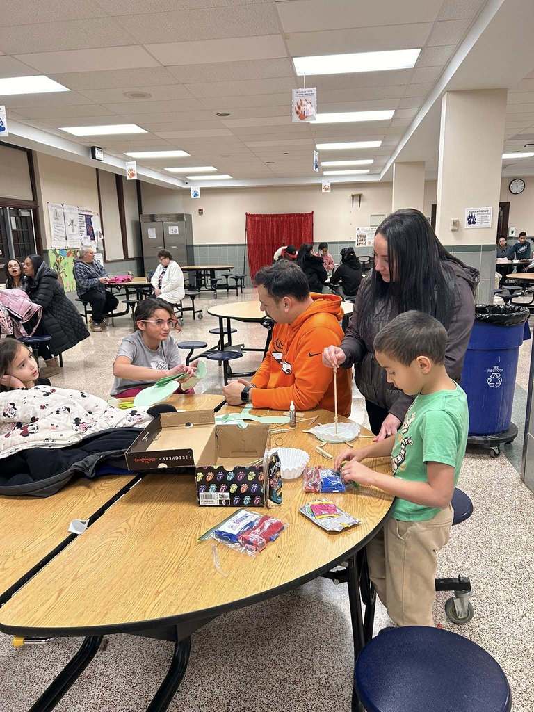 Two adults and three children are sitting at a round table in a school cafeteria participating in a craft activity. Other adults and children can be seen sitting at round tables in the background.