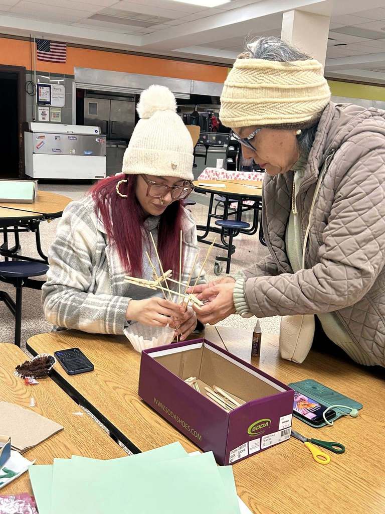 Two adult women are gathered at a round table working together on a craft project using clothespins in a school cafeteria. 