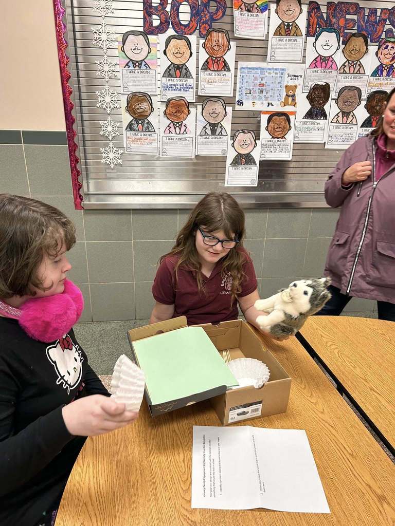Two children are sitting at a round table in a school cafeteria, engaging in a craft project. An adult and a bulletin board with children's art can be seen nearby in the background.