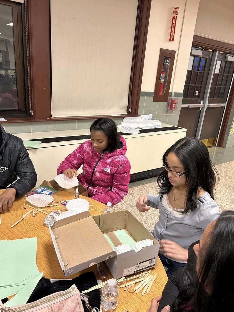 Two young girls are sitting at a round table in a school cafeteria doing crafts with coffee filters and wood sticks. Other people can be seen sitting at the table.