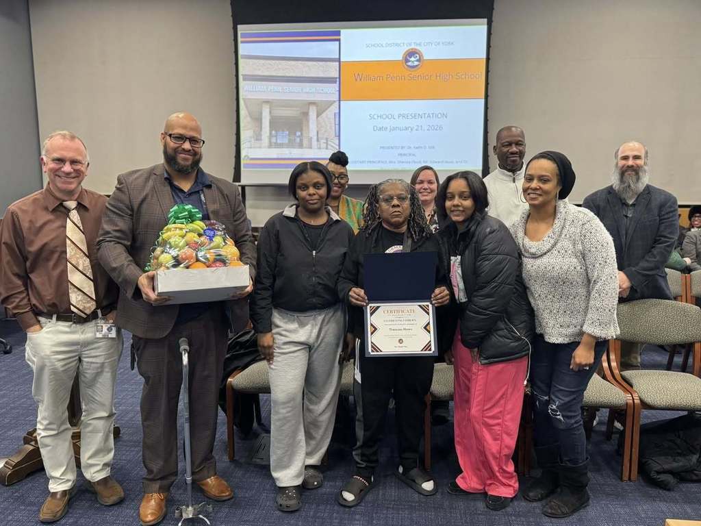 A group of people, including including administration, a district staff member, and a district family are standing in a conference room. One administrator is holding a gift basket, while a district parent is holding a certificate. A presentation slide is displayed on a screen in the background.