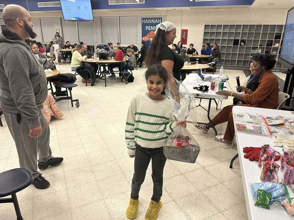 A young girl is standing in a school cafeteria, holding a wrapped gift basket. Other people can be seen standing and sitting at round tables in the background.