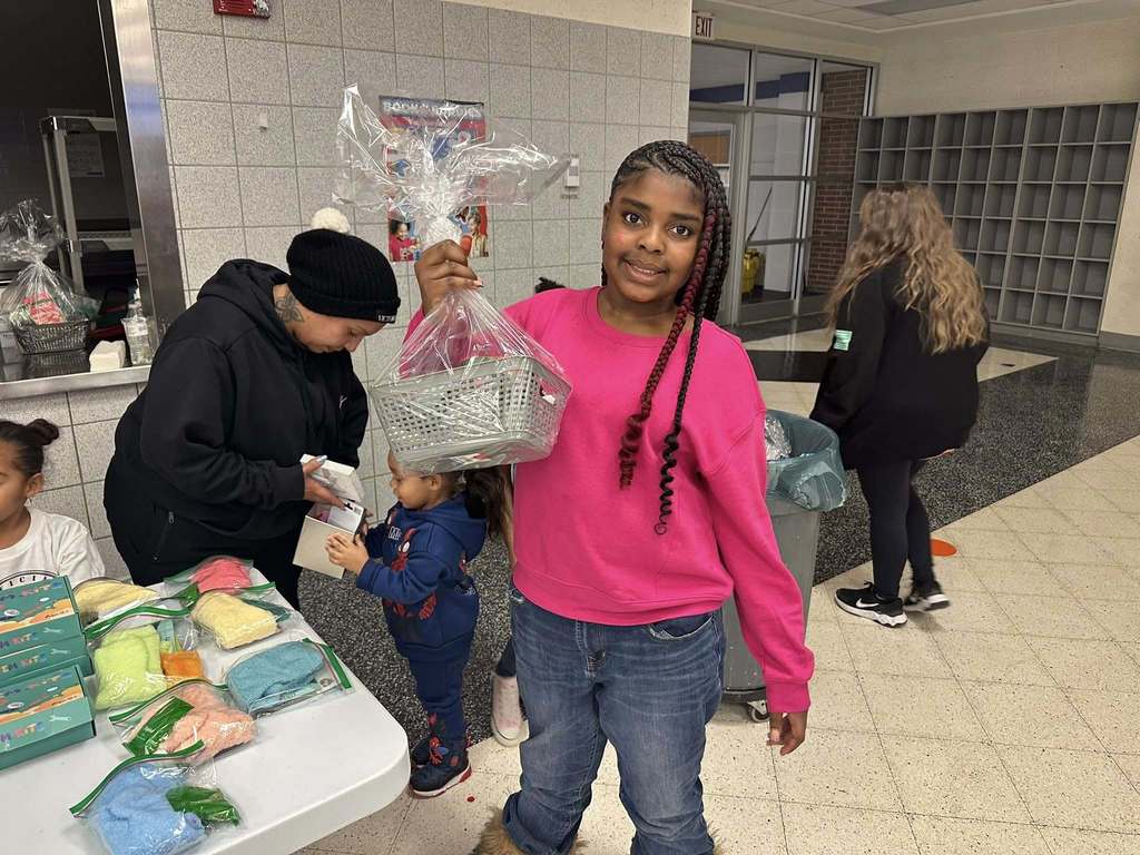 A young girl is holding up a wrapped gift basket in a school cafeteria. Other people can be seen in the background.