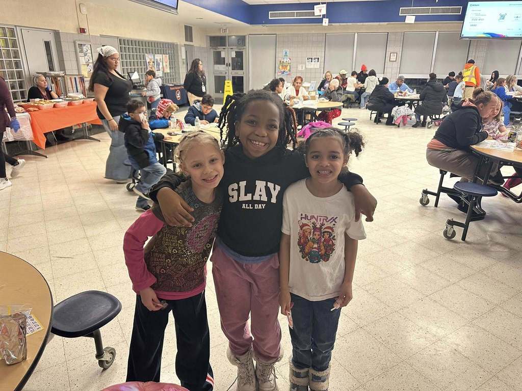 A group of three young girls are standing together in a school cafeteria. One of the girls has her arms wrapped around the other two girls. Other people can be seen in the background.
