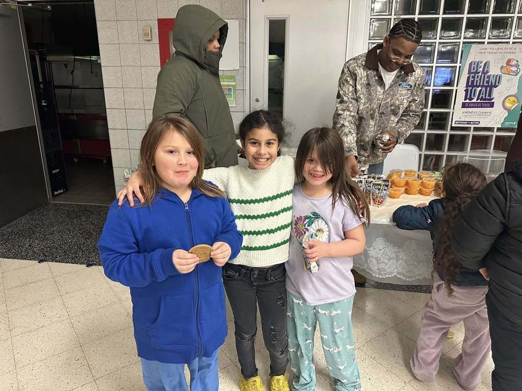A group of three young girls are standing together in a school cafeteria, with one of them holding a cookie and another holding a capri sun . One of the girls has her arm wrapped around the other two girls. A person can be seen serving food at a table in the background.