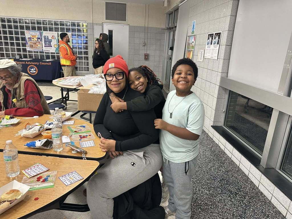 A woman is sitting at a round table in school cafeteria and two young children are nearby, with one of them having her arms wrapped around the woman. Other people can be seen in the background.